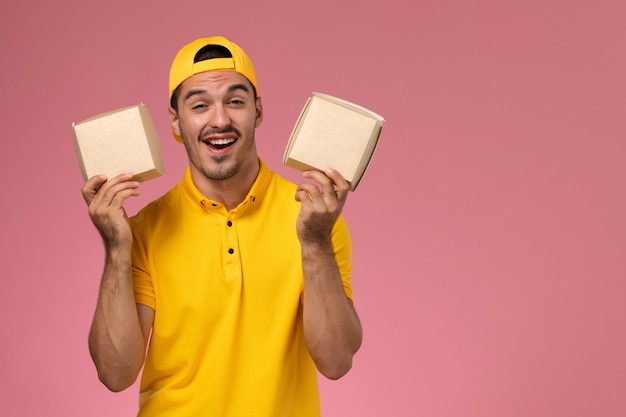 Mensajero masculino de vista frontal en uniforme amarillo sosteniendo pequeños paquetes de alimentos sobre fondo rosa.