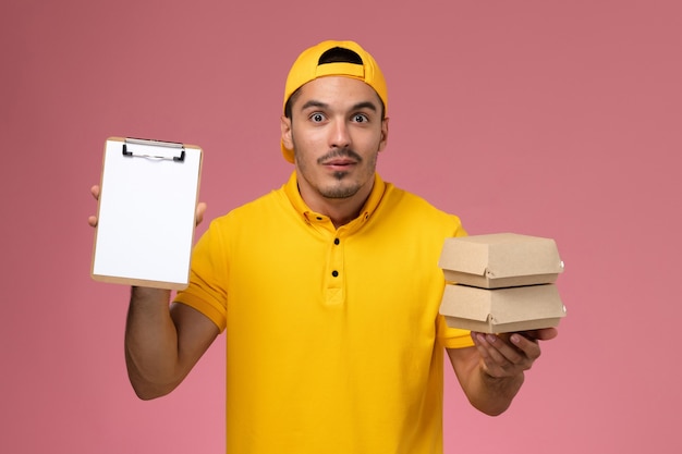 Mensajero masculino de vista frontal en uniforme amarillo sosteniendo pequeños paquetes de comida y un pequeño bloc de notas sobre fondo rosa claro.
