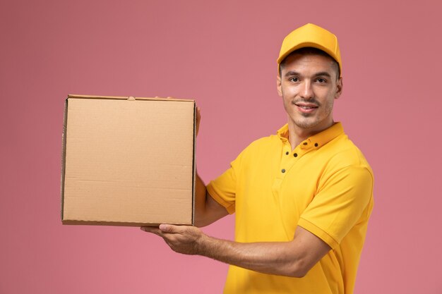 Mensajero masculino de vista frontal en uniforme amarillo sosteniendo posign de caja de entrega de alimentos con él en el fondo rosa