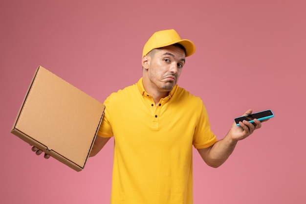 Mensajero masculino de vista frontal en uniforme amarillo sosteniendo su teléfono y caja de entrega de alimentos en el escritorio rosa