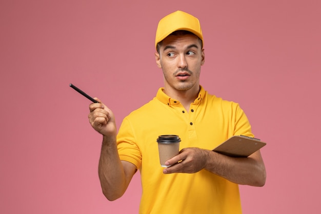 Mensajero masculino de vista frontal en uniforme amarillo sosteniendo la taza de café de entrega y el bloc de notas escribiendo notas en el escritorio rosa claro