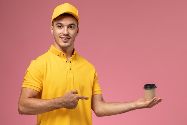 Mensajero masculino de vista frontal en uniforme amarillo sosteniendo la taza de entrega de café en el fondo rosa