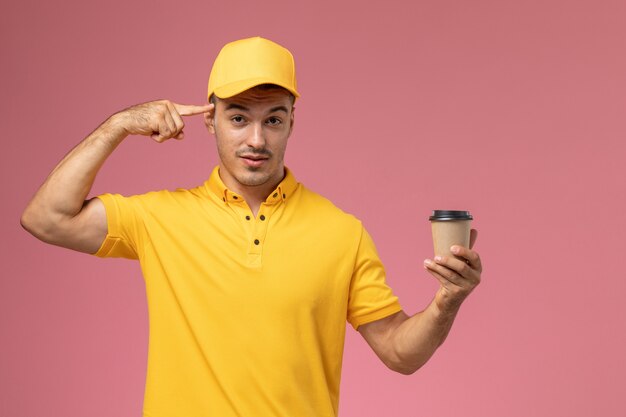 Mensajero masculino de vista frontal en uniforme amarillo sosteniendo la taza de entrega de café marrón tocando su sien en el escritorio rosa