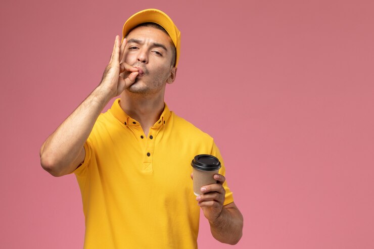 Foto gratuito mensajero masculino de vista frontal en uniforme amarillo sosteniendo la taza de entrega de café que muestra sabroso cartel en el escritorio rosa