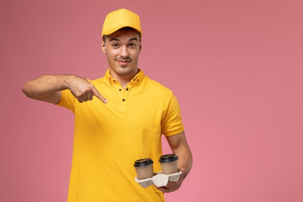Mensajero masculino de vista frontal en uniforme amarillo sosteniendo tazas de café de entrega en el escritorio de color rosa claro