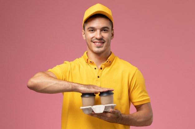 Mensajero masculino de vista frontal en uniforme amarillo sosteniendo tazas de café de entrega en escritorio rosa