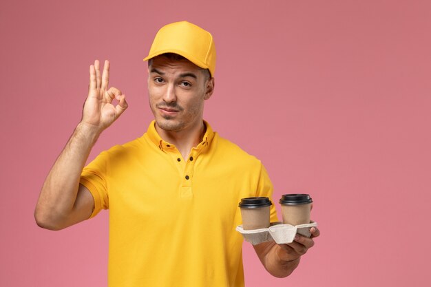 Mensajero masculino de vista frontal en uniforme amarillo sosteniendo tazas de café de entrega en el fondo rosa claro