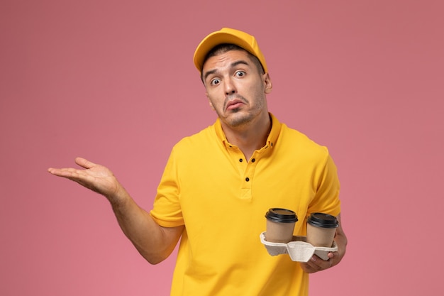 Mensajero masculino de vista frontal en uniforme amarillo sosteniendo tazas de café de entrega en el fondo rosa claro