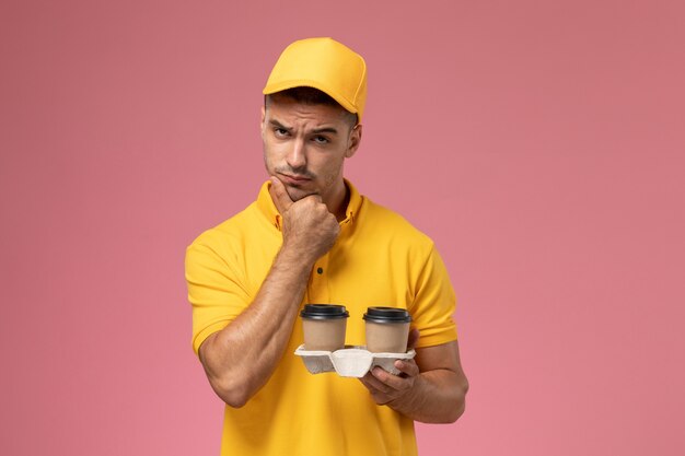 Mensajero masculino de vista frontal en uniforme amarillo sosteniendo tazas de café de entrega en el fondo rosa claro