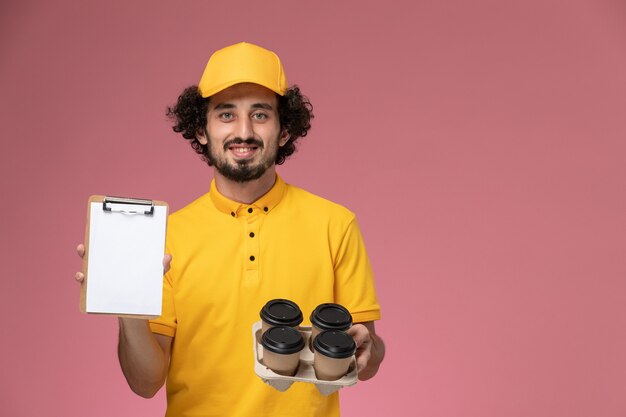 Mensajero masculino de vista frontal en uniforme amarillo sosteniendo tazas de café de entrega marrón y bloc de notas en la pared rosa
