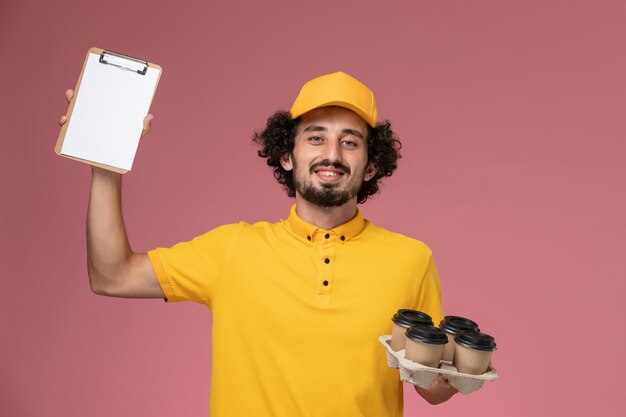 Mensajero masculino de vista frontal en uniforme amarillo sosteniendo tazas de café de entrega marrón y bloc de notas en la pared rosa