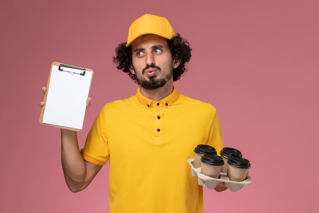 Mensajero masculino de vista frontal en uniforme amarillo sosteniendo tazas de café de entrega marrón y bloc de notas en la pared rosa