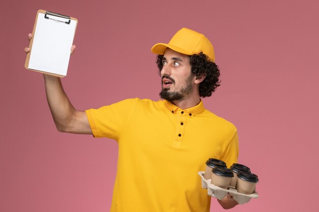 Mensajero masculino de vista frontal en uniforme amarillo sosteniendo tazas de café de entrega marrón y bloc de notas en la pared rosa