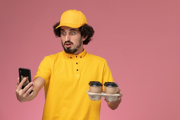 Mensajero masculino de vista frontal en uniforme amarillo sosteniendo tazas de café de entrega marrón hablando por teléfono en la pared rosa