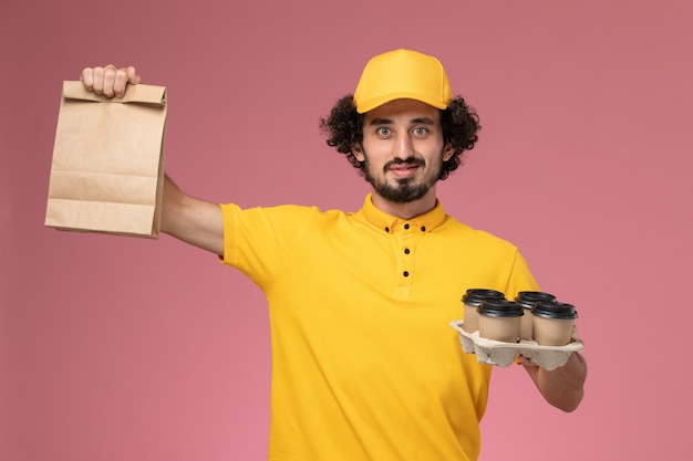 Mensajero masculino de vista frontal en uniforme amarillo sosteniendo tazas de café de entrega marrón y paquete de comida en la pared rosa