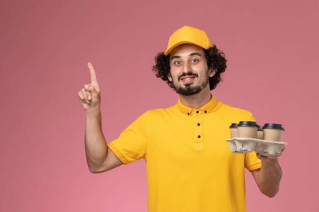 Mensajero masculino de vista frontal en uniforme amarillo sosteniendo tazas de café de entrega marrón en la pared rosa