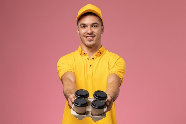 Mensajero masculino de vista frontal en uniforme amarillo sosteniendo tazas de café de entrega y sonriendo en el escritorio rosa