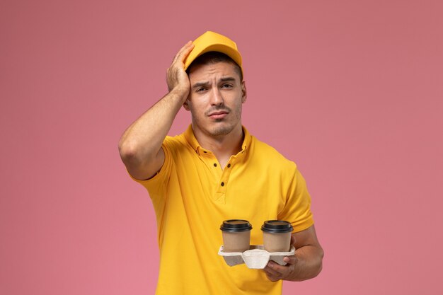 Mensajero masculino de vista frontal en uniforme amarillo sosteniendo tazas de café de entrega sosteniendo su cabeza sobre fondo rosa claro