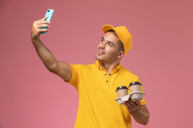 Mensajero masculino de vista frontal en uniforme amarillo sosteniendo tazas de café de entrega tomando selfie en escritorio rosa