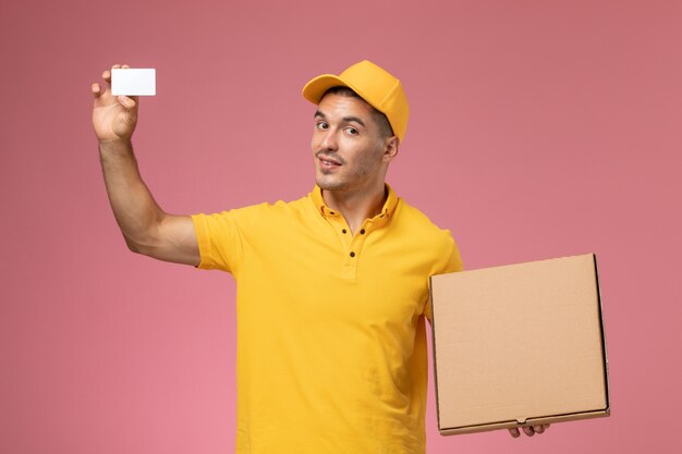 Mensajero masculino de vista frontal en uniforme amarillo con tarjeta blanca y caja de entrega de alimentos en el escritorio rosa