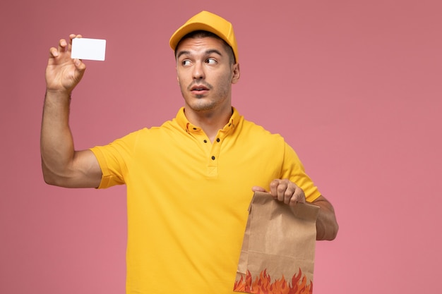 Mensajero masculino de vista frontal en uniforme amarillo con tarjeta blanca y paquete de alimentos en el escritorio rosa