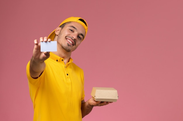 Mensajero masculino de vista frontal en uniforme amarillo con tarjeta y pequeño paquete de comida sobre fondo rosa.