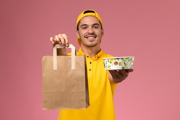 Mensajero masculino de vista frontal en uniforme amarillo con tazón de paquete de comida de entrega sonriendo sobre fondo rosa.