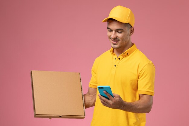 Mensajero masculino de vista frontal en uniforme amarillo con teléfono y sosteniendo la caja de entrega de alimentos en el escritorio rosa