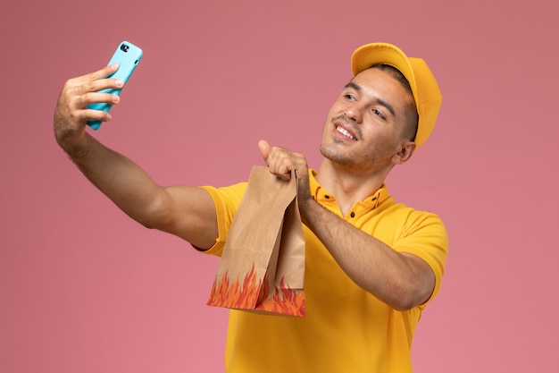 Mensajero masculino de vista frontal en uniforme amarillo tomando una foto con paquete de alimentos en el escritorio rosa