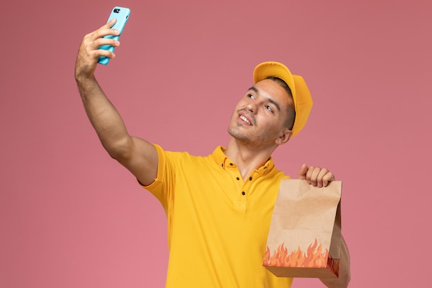 Mensajero masculino de vista frontal en uniforme amarillo tomando un selfie con paquete de comida en el escritorio rosa