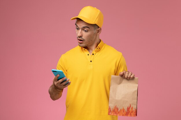 Mensajero masculino de vista frontal en uniforme amarillo usando su teléfono sosteniendo el paquete de alimentos en el escritorio rosa