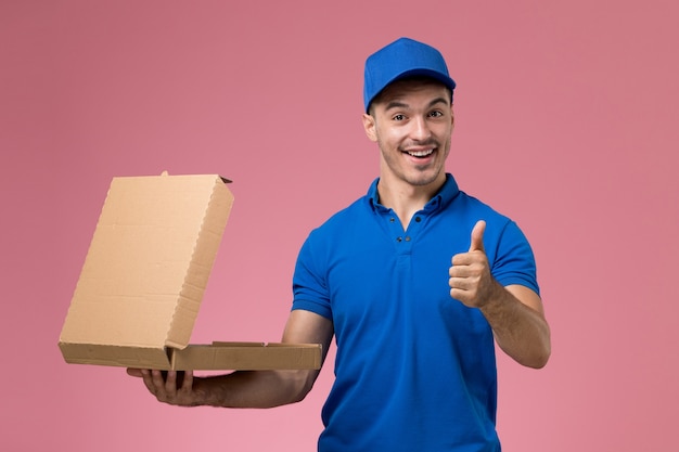 Mensajero masculino de vista frontal en uniforme azul con caja de comida sonriendo en la pared rosa, entrega de servicio uniforme de trabajador de trabajo