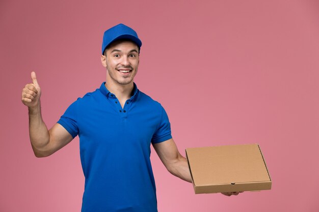 Mensajero masculino de vista frontal en uniforme azul con caja de comida con sonrisa en la pared rosa, entrega de trabajo de servicio uniforme