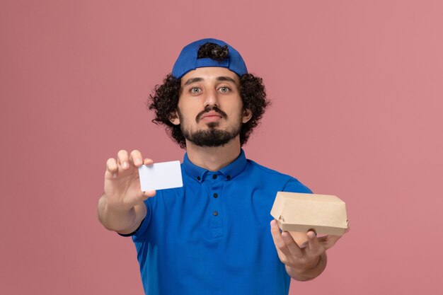 Mensajero masculino de vista frontal en uniforme azul y capa con paquete de comida de entrega y tarjeta en trabajo de servicio de entrega uniforme de pared rosa