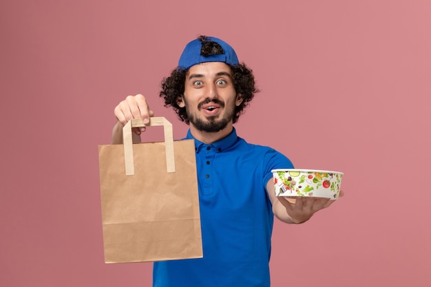 Foto gratuita mensajero masculino de vista frontal en uniforme azul y capa con paquete de comida de entrega y tazón en la pared rosa uniforme de servicio de entrega masculino