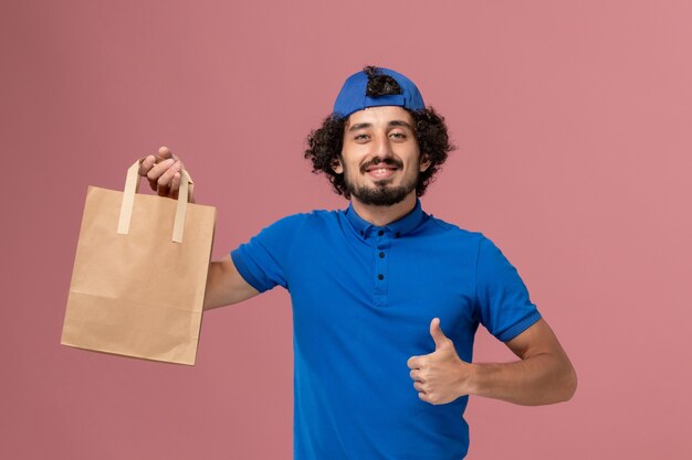 Mensajero masculino de vista frontal en uniforme azul y capa con paquete de comida de papel de entrega en el trabajador uniforme de servicio de entrega de pared rosa
