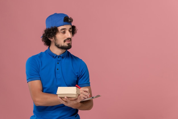 Mensajero masculino de vista frontal en uniforme azul y capa con pequeño paquete de comida de entrega y bloc de notas escribiendo en servicio de entrega uniforme de pared rosa