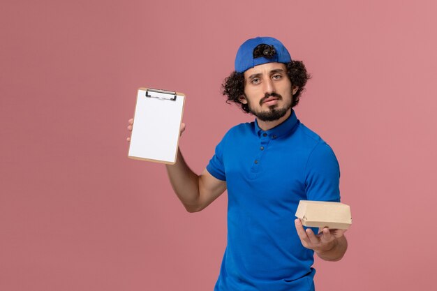 Mensajero masculino de vista frontal en uniforme azul y capa con pequeño paquete de comida de entrega y bloc de notas en la pared rosa trabajador de servicio de entrega uniforme