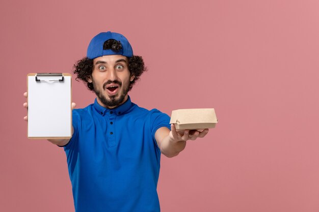 Mensajero masculino de vista frontal en uniforme azul y capa con pequeño paquete de comida de entrega y bloc de notas en servicio de trabajo de entrega uniforme de pared rosa