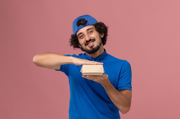 Mensajero masculino de vista frontal en uniforme azul y capa con pequeño paquete de comida de entrega sonriendo en la pared rosa