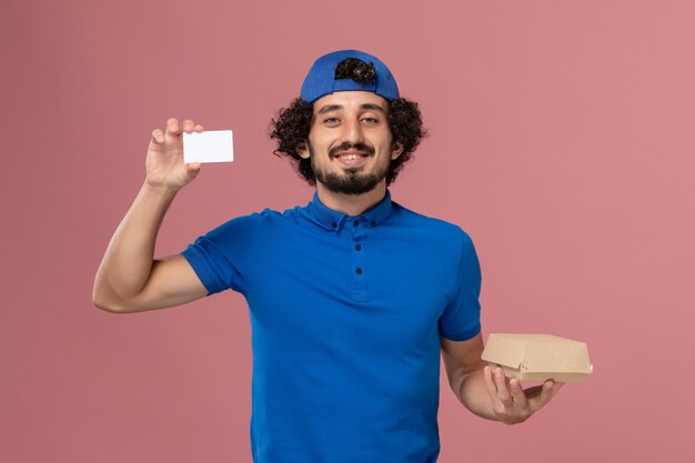 Mensajero masculino de vista frontal en uniforme azul y capa con pequeño paquete de comida de entrega y tarjeta en el servicio de entrega uniforme de pared rosa