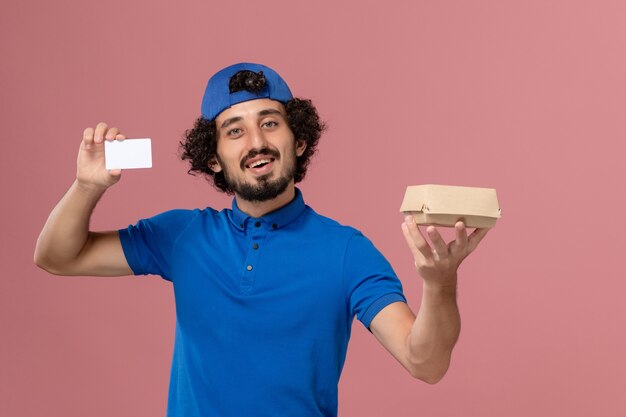 Mensajero masculino de vista frontal en uniforme azul y capa con pequeño paquete de comida de entrega y tarjeta en servicio de trabajador de entrega uniforme de pared rosa