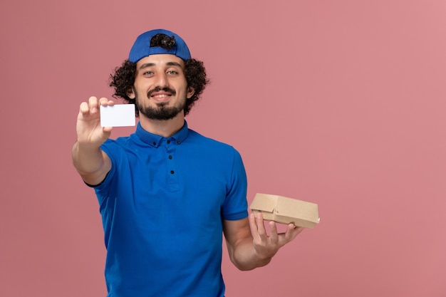 Mensajero masculino de vista frontal en uniforme azul y capa con pequeño paquete de comida de entrega y tarjeta en servicio de trabajo de entrega uniforme de pared rosa