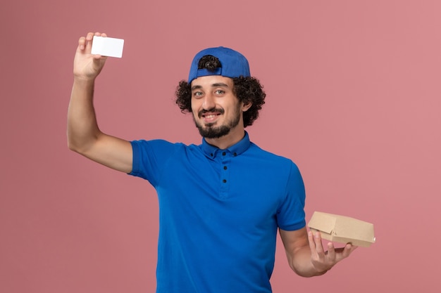 Mensajero masculino de vista frontal en uniforme azul y capa con pequeño paquete de comida de entrega y tarjeta en servicio de trabajo de entrega uniforme de pared rosa
