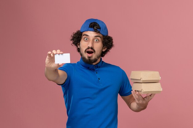 Mensajero masculino de vista frontal en uniforme azul y capa sosteniendo paquetes de comida de entrega con tarjeta en el trabajo uniforme de servicio de entrega de pared rosa