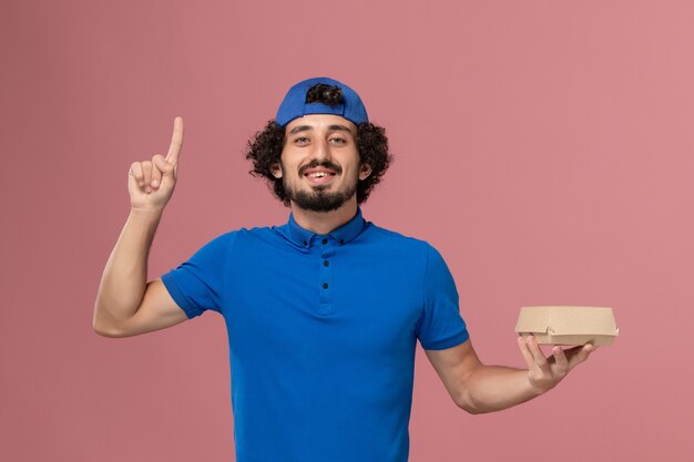 Mensajero masculino de vista frontal en uniforme azul y capa sosteniendo un pequeño paquete de comida de entrega con el dedo levantado en la pared rosa