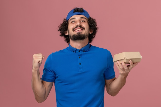 Mensajero masculino de vista frontal en uniforme azul y capa sosteniendo un pequeño paquete de comida de entrega en la pared rosa