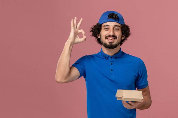 Mensajero masculino de vista frontal en uniforme azul y capa sosteniendo un pequeño paquete de comida de entrega en la pared rosa