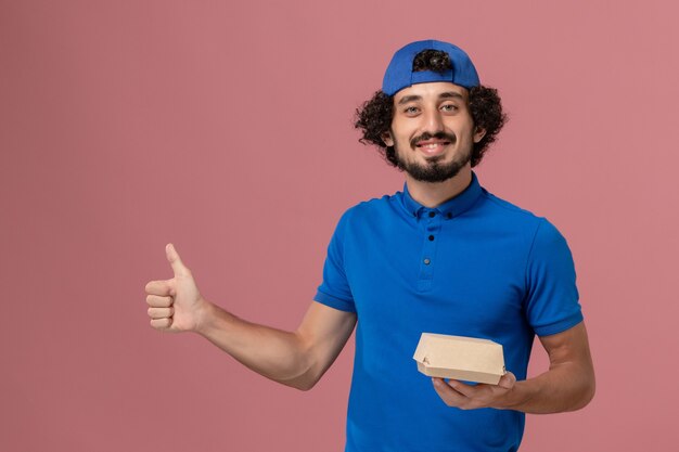 Mensajero masculino de vista frontal en uniforme azul y capa sosteniendo un pequeño paquete de comida de entrega en la pared rosa