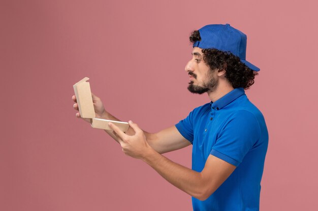 Mensajero masculino de vista frontal en uniforme azul y capa sosteniendo un pequeño paquete de comida de entrega en la pared rosa